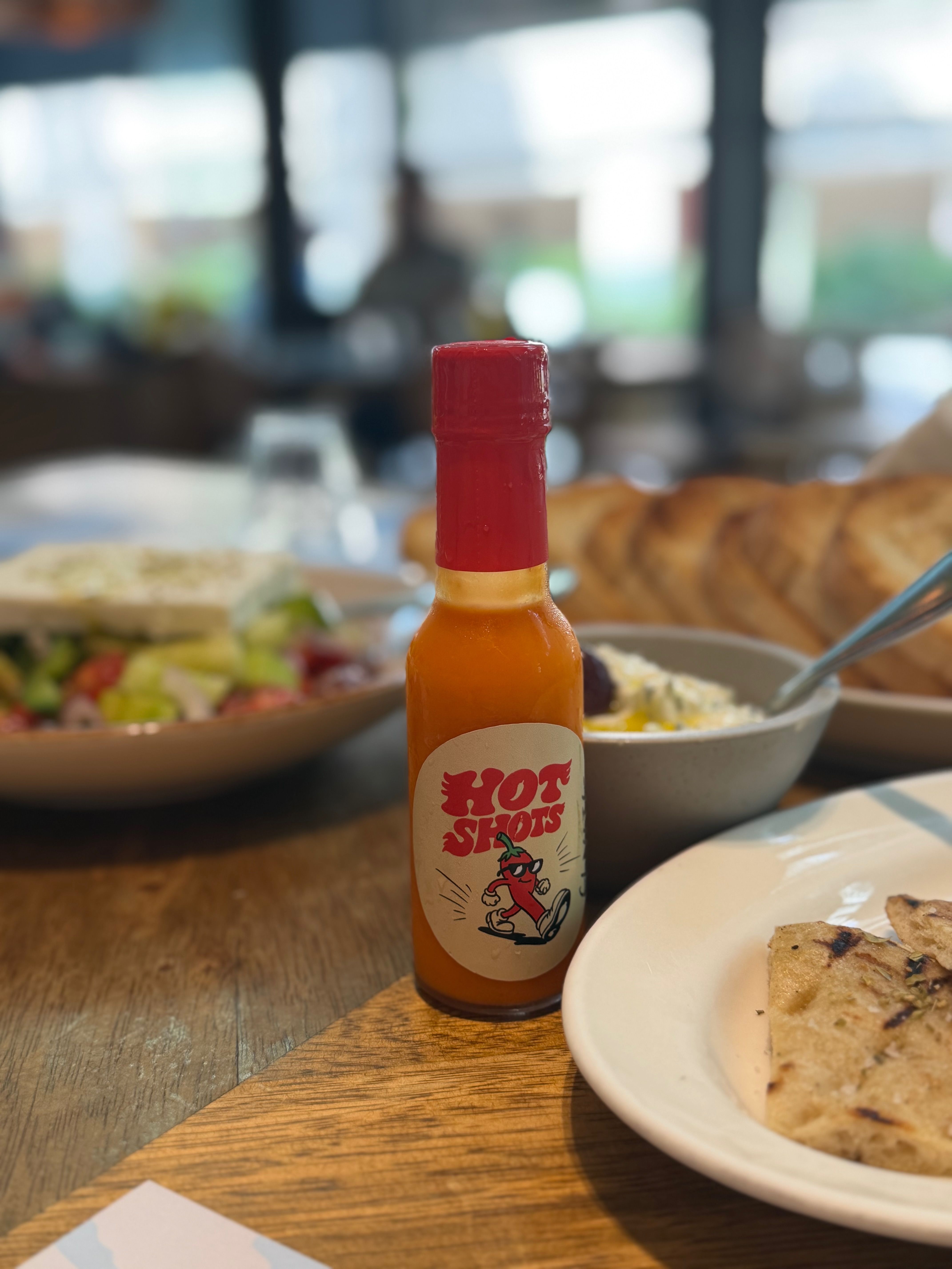 A bottle of Hot Shots on a café table beside grilled bread and a shared plate