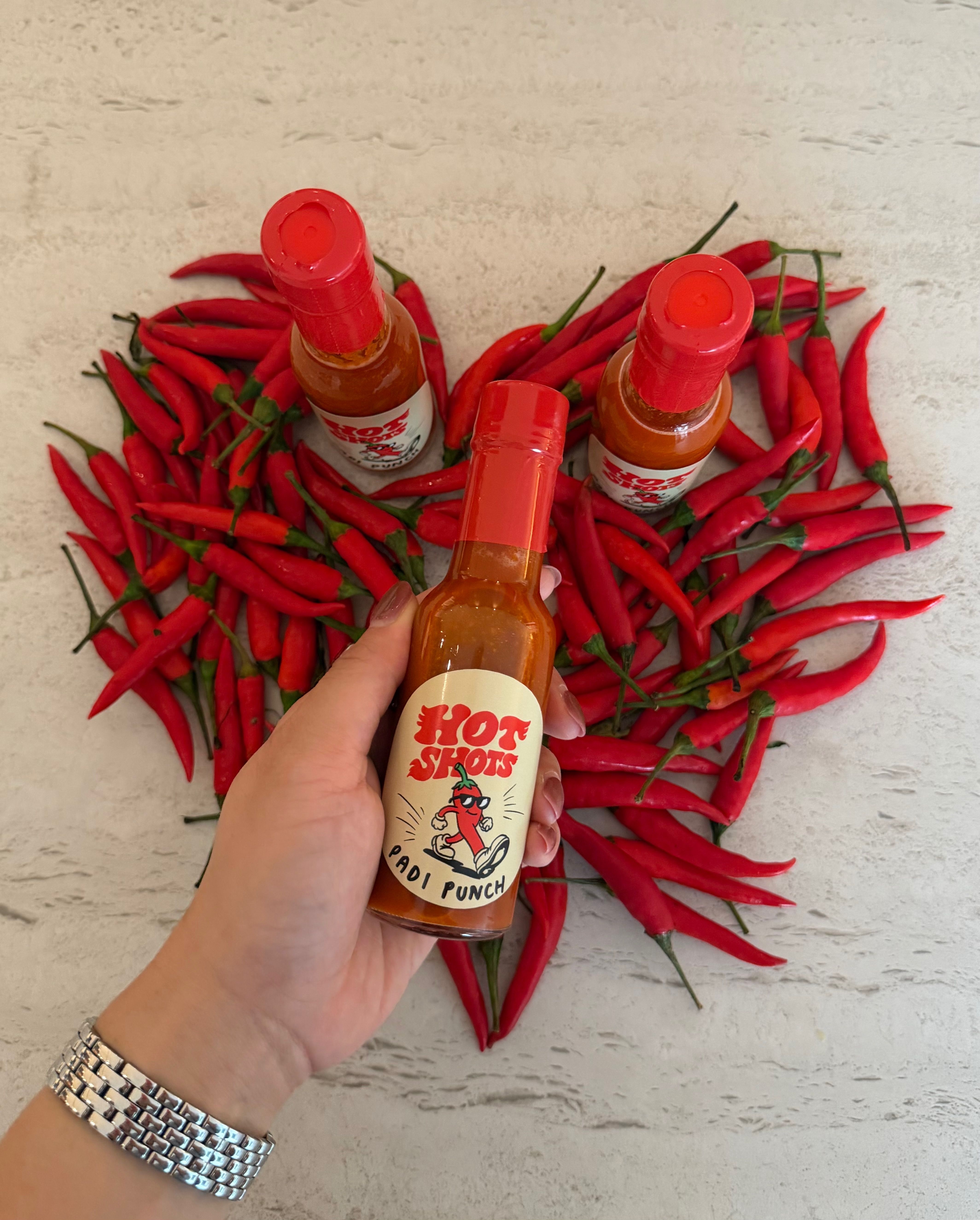 A hand holding a bottle of Hot Shots Padi Punch over chilli padi arranged in a heart shape